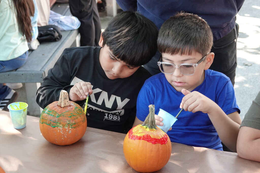 Two elementary school boys sit side-by-side at a outdoor paper-covered table, focused on decorating pumpkins. The boy on the left uses a paintbrush to apply green paint in rings around his pumpkin, while the boy on the right paints red and yellow details near the stem of his pumpkin.