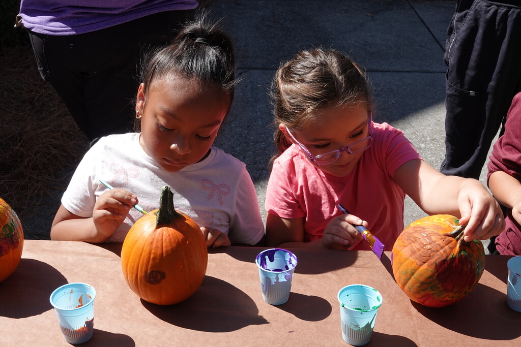 Two elementary school girls sit next to each other, looking down attentively as they decorate pumpkins with paintbrushes. Small plastic cups holding colorful paint sit on the craft table in front of them.