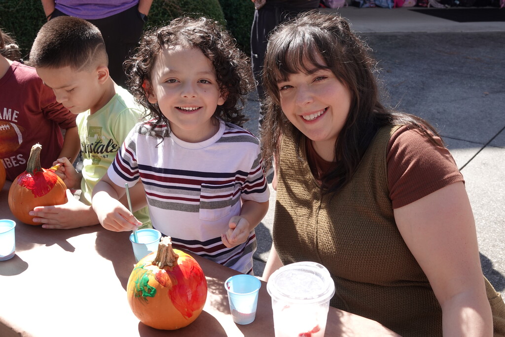 A young boy in a striped shirt and a smiling adult female educator pose together at an outdoor table. In front of them are small pumpkins decorated with colorful paint and cups of water with paintbrushes.