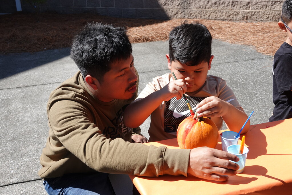 An adult man in an olive long-sleeve shirt leans in close to assist a young boy as he carefully paints red stripes onto a small pumpkin. Small plastic cups filled with paint and water sit on the orange table in front of them.