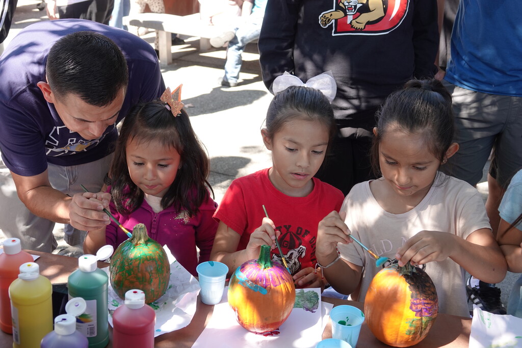 Three elementary school girls sit at a table painting small pumpkins in vivid green, purple, yellow, and orange patterns. On the left, an adult man leans down to guide one girl's brush, while bottles of colorful craft paint sit on the table.