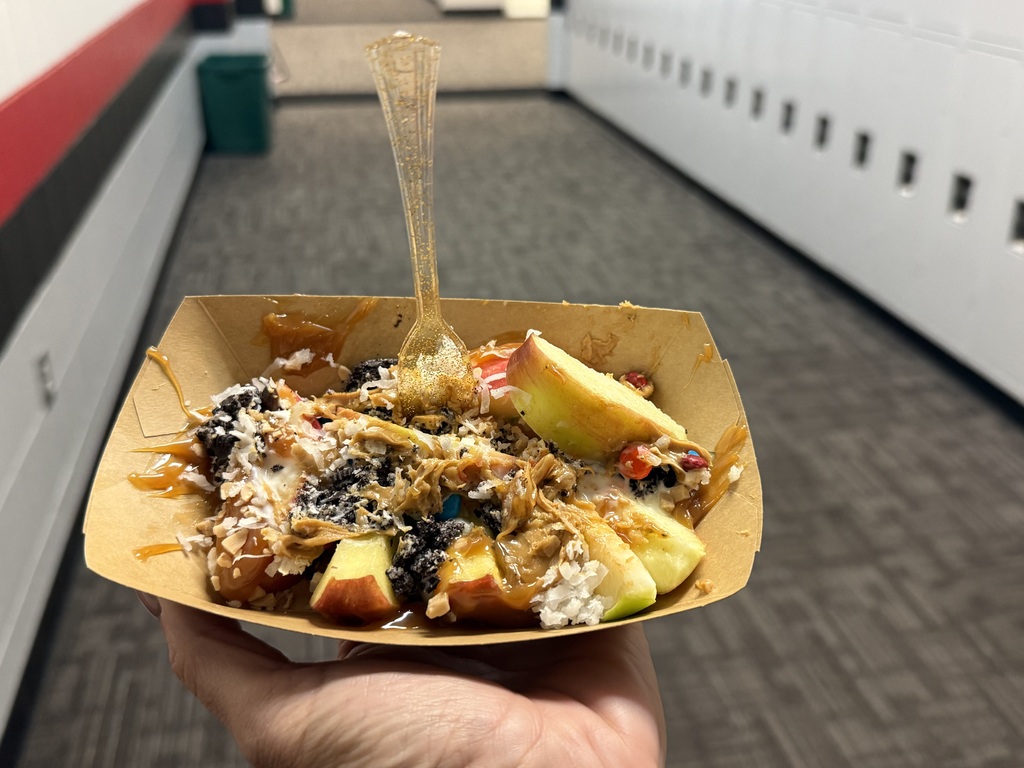 A cardboard food boat filled with sliced apples topped with caramel drizzle, peanut butter, crushed Oreo cookies, coconut, and M&Ms, held in someone's hand.