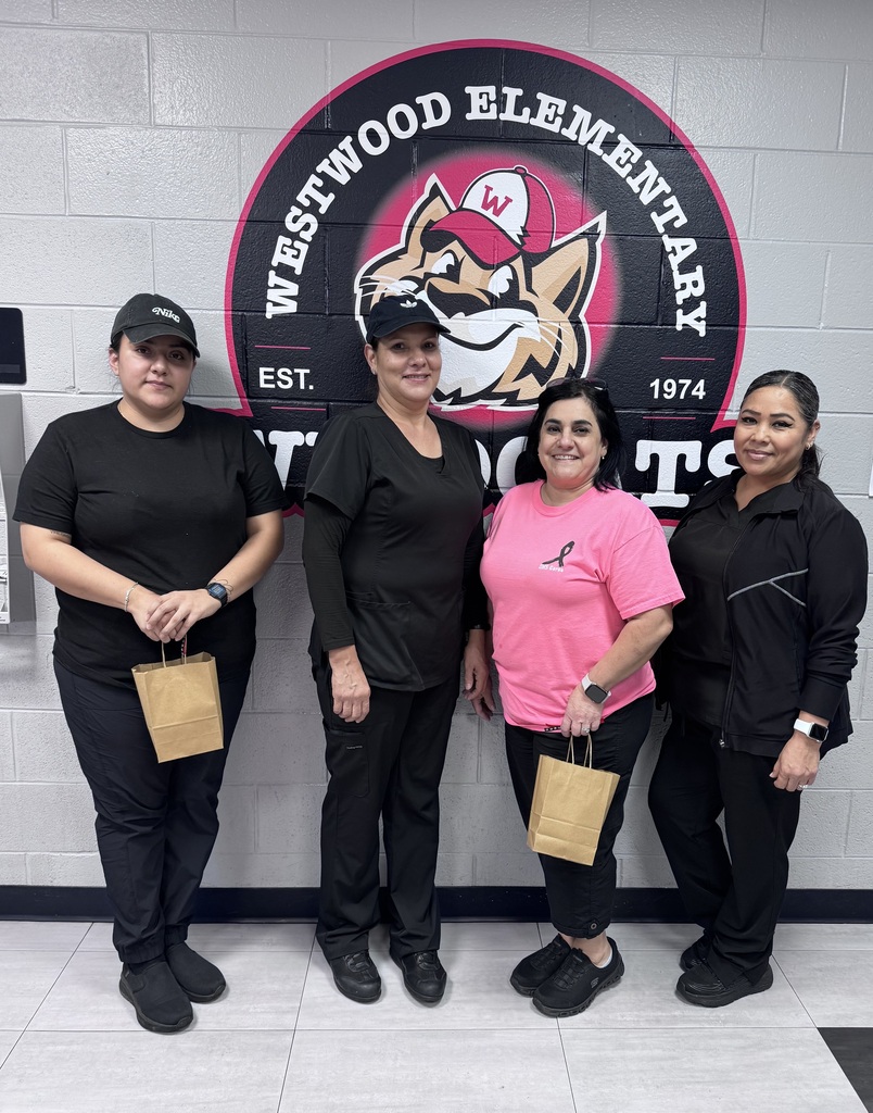 Four women stand in a row in front of a large circular Westwood Elementary logo painted on a cinderblock wall. Three of the women wear black outfits and black caps or jackets, while one woman wears a pink t-shirt featuring a breast cancer awareness ribbon. Two of the women hold small, brown paper gift bags.