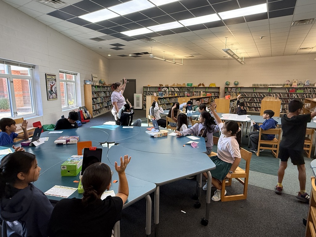 A group of elementary students sit around large, curved blue tables in the school media center, raising their hands to participate in a lesson led by a teacher standing near the bookshelves in the background.