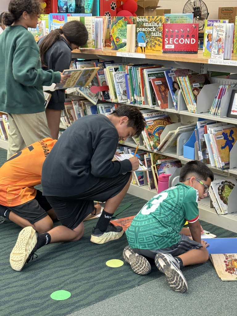 Several elementary school students gather in a library aisle in front of colorful book stacks. Two boys kneel on the carpet floor writing on clipboards, while other classmates stand and crouch nearby browsing books on lower shelves.