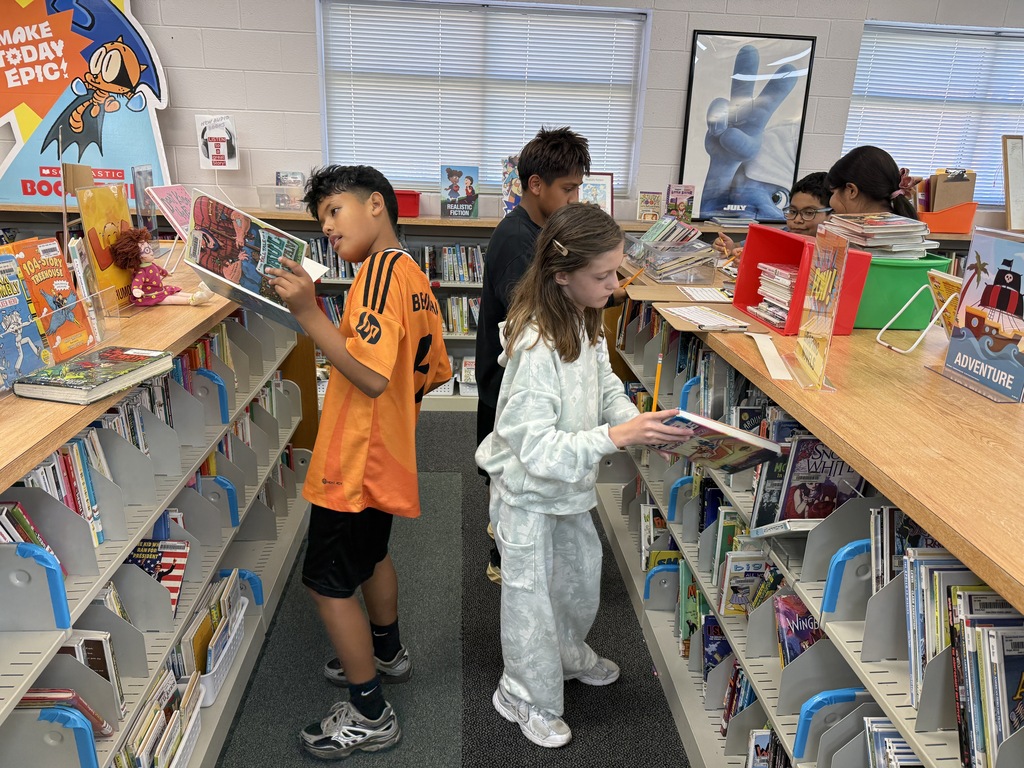 Elementary school students stand between rows of library book stacks, attentively reading and browsing through picture books and graphic novels displayed on top of low shelves.