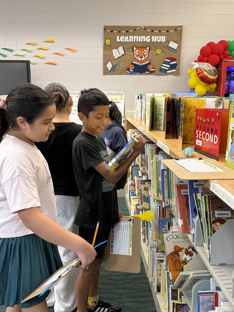 Students holding clipboards and browsing bookshelves during a library scavenger hunt.