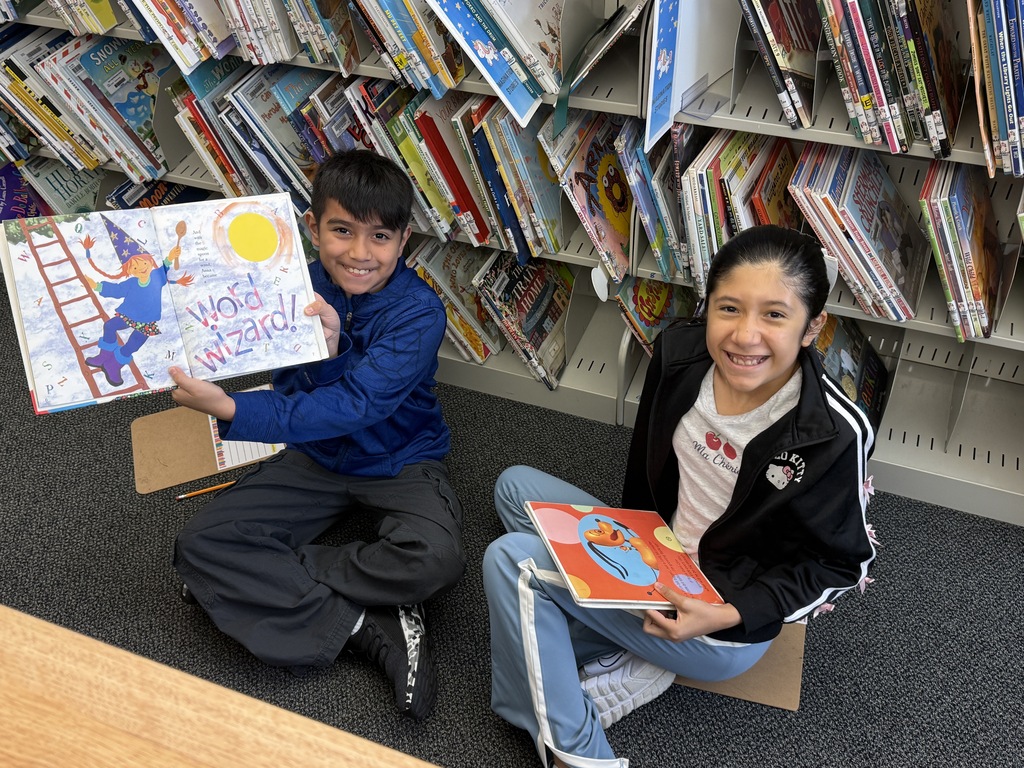 Two students sitting on the library floor holding open children's books and smiling.