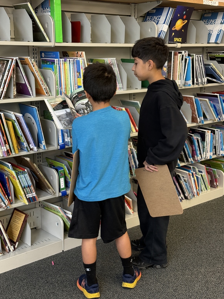 Two young boys stand together in front of library book stacks. The boy in the bright blue shirt holds open a book about World War II, showing it to his classmate in a black hoodie who holds a clipboard.