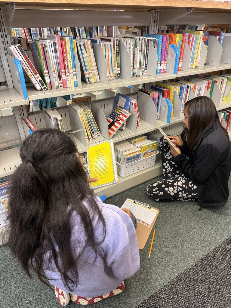 Two elementary school girls sitting on the library carpet reading books with clipboards nearby.