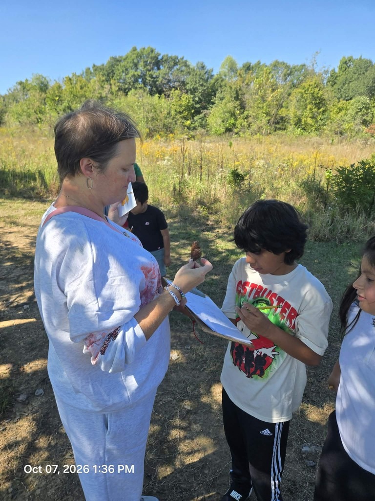 A female educator stands outdoors holding a small natural specimen, talking with a boy in a graphic t-shirt as he writes on a clipboard worksheet.