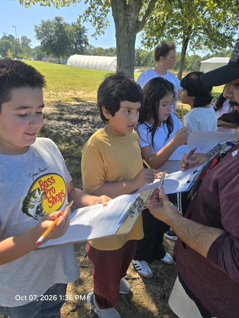 A group of students gather outdoors in a grassy area, taking notes on clipboard worksheets while an instructor holds up plant leaves for close inspection.