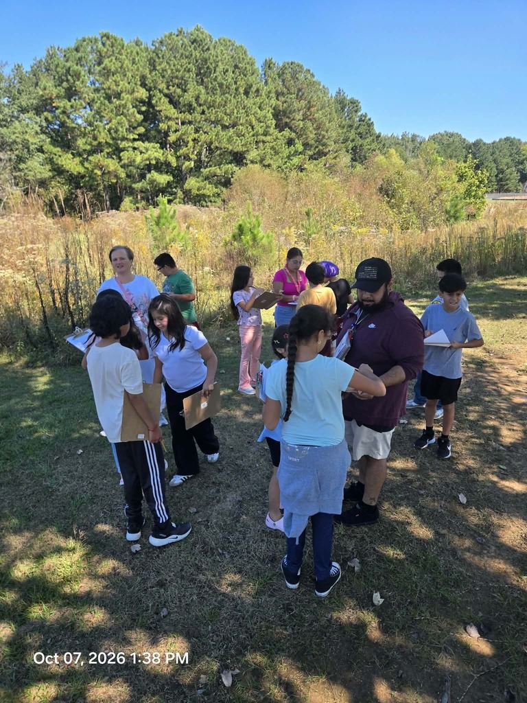 A group of elementary students stand in a sunlit field near tall grass and trees, working on clipboards while talking with teachers during an outdoor science lesson.