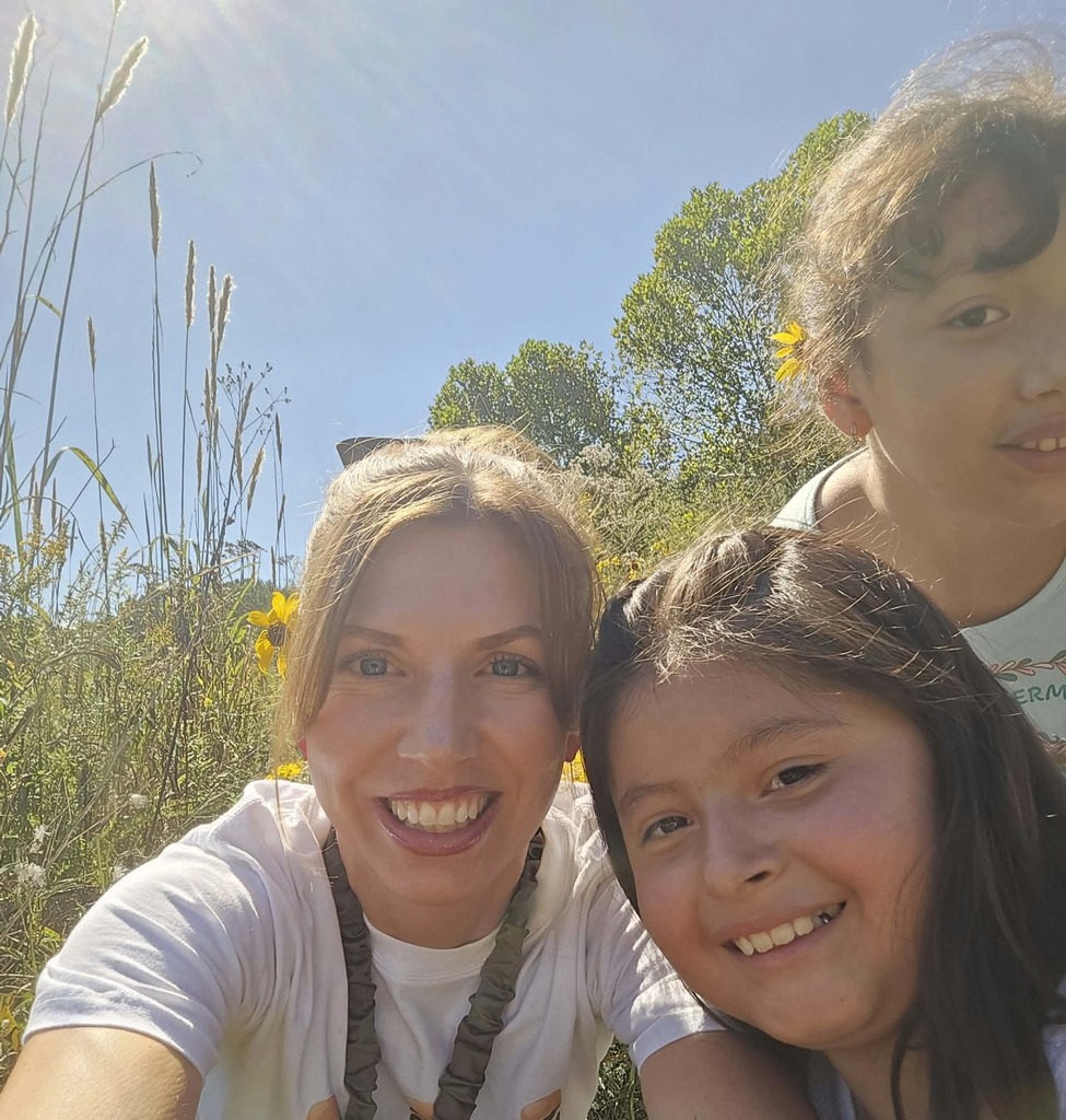 A close-up outdoor selfie of a smiling female teacher standing alongside two young female students, with small yellow wildflowers tucked behind their ears against a bright blue sky.