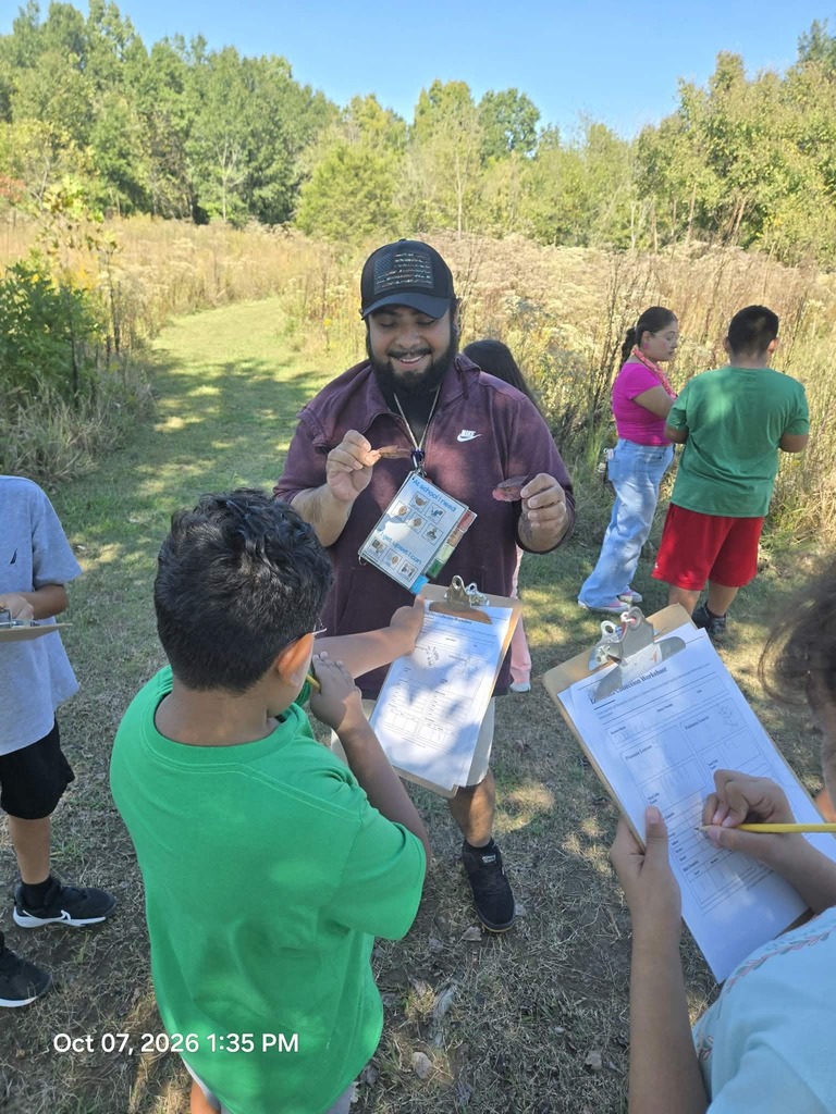 An instructor in a cap holds up dried leaves for students in a field setting as they fill out data collection worksheets on clipboards.