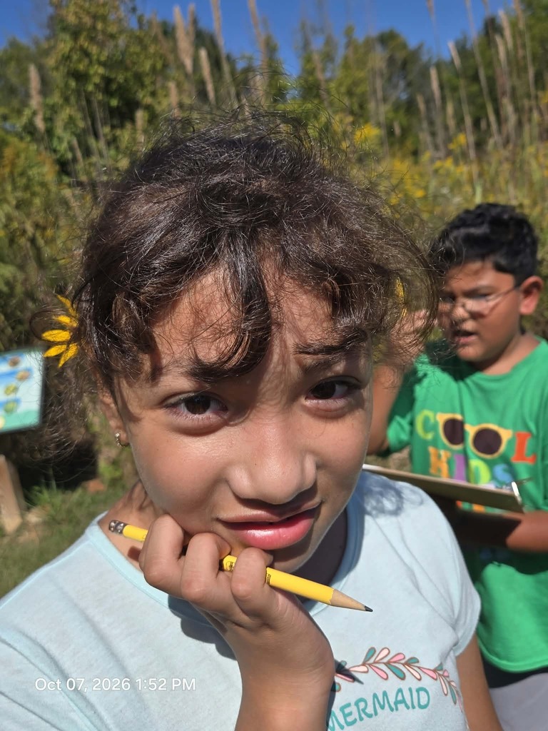A close-up portrait of a young girl with a yellow wildflower tucked behind her ear, leaning her chin on a pencil while looking into the camera. In the background, a boy in a green shirt holds a clipboard during an outdoor learning activity in a grassy field.