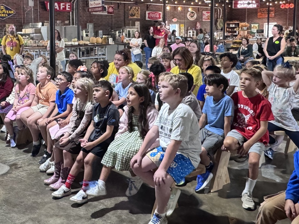 Students and parents smiling while riding on a wooden flatbed trailer pulled by a tractor during an outdoor farm field trip. The group sits along the green wooden side rails under a bright blue sky.