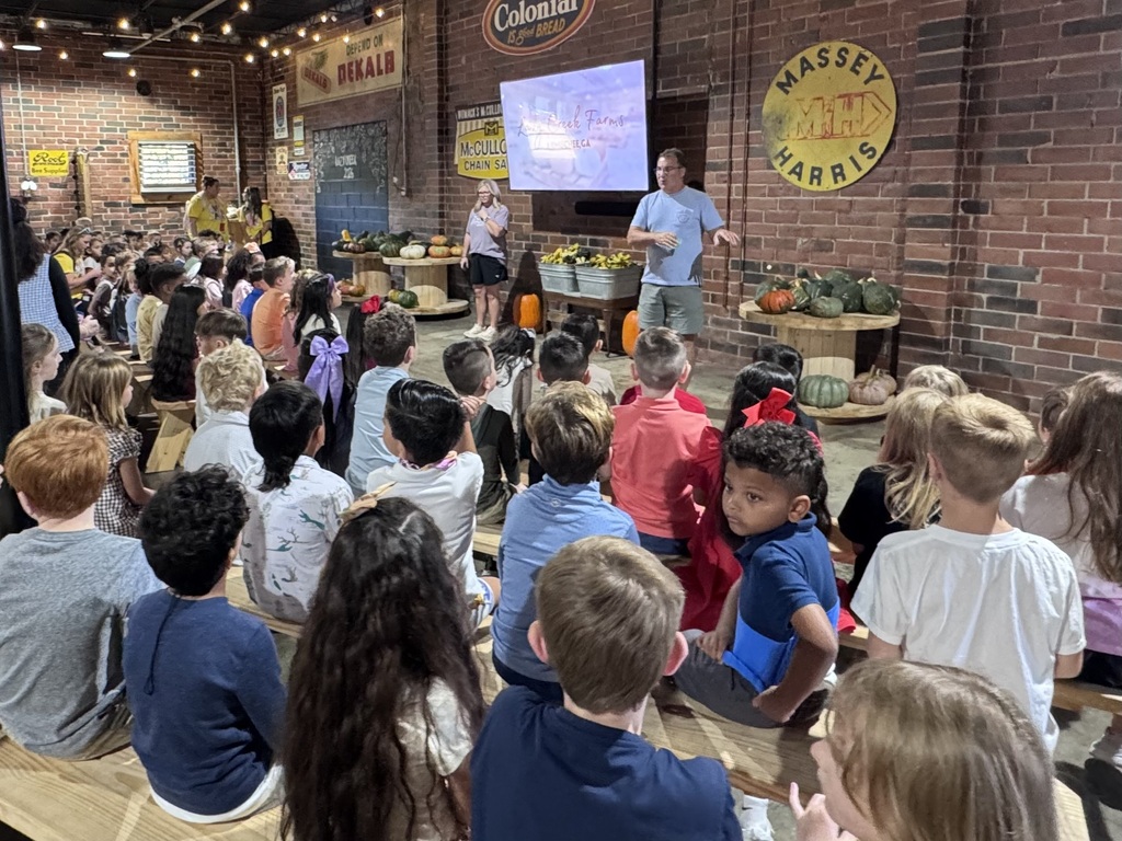 Students sit on wooden benches in a rustic brick-walled room with vintage signs and displays of pumpkins and gourds. At the front of the room, two speakers give a presentation in front of a projection screen.