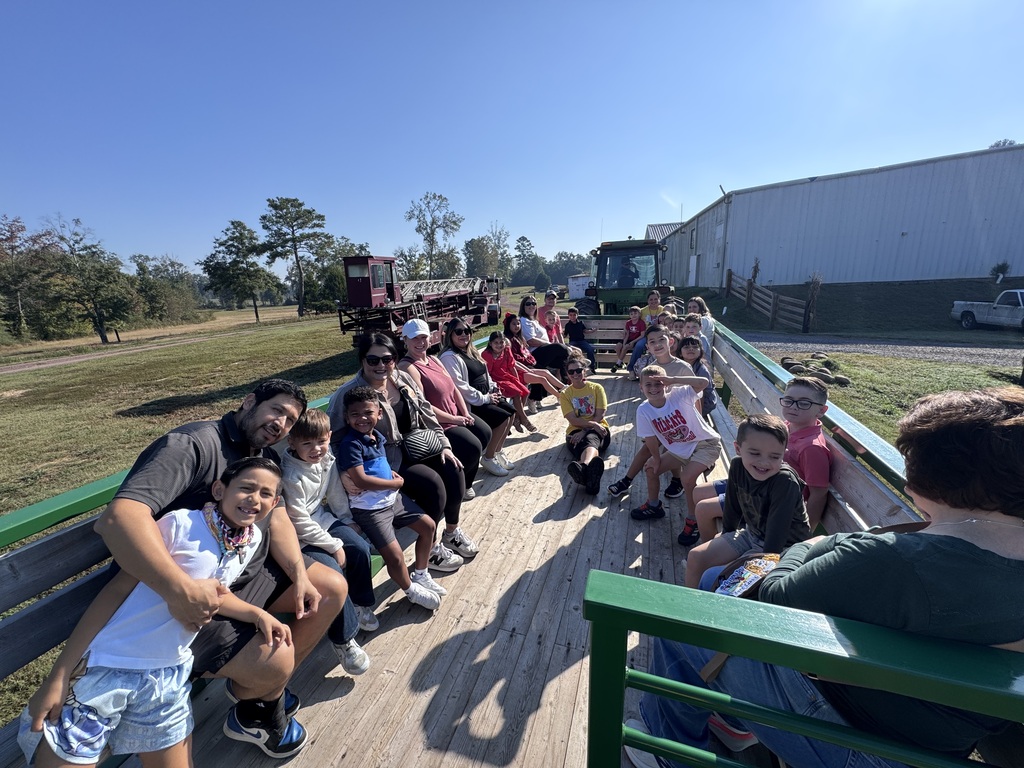 Students and parents smiling while riding on a wooden flatbed trailer pulled by a tractor during an outdoor farm field trip. The group sits along the green wooden side rails under a bright blue sky.