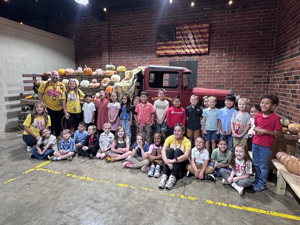 Elementary students and teachers gather together for a group picture indoors. Behind them, a vintage red truck and wooden crates display an assortment of pumpkins and gourds against a brick wall decorated with a wooden American flag.