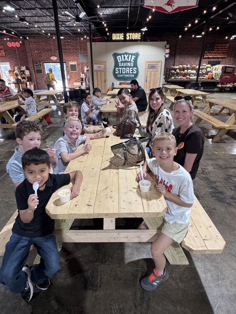 Students and parents smile for a photo while seated at a long wooden picnic table enjoying snacks in cups. The indoor venue features string lights, brick walls, and vintage store signage in the background.