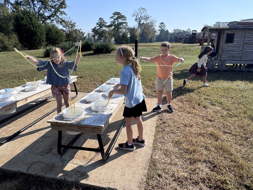 Elementary children standing around outdoor wooden tables making large bubbles with wands and bowls of soap solution. Green grass, trees, and a wooden play structure are visible in the background under a clear sky.