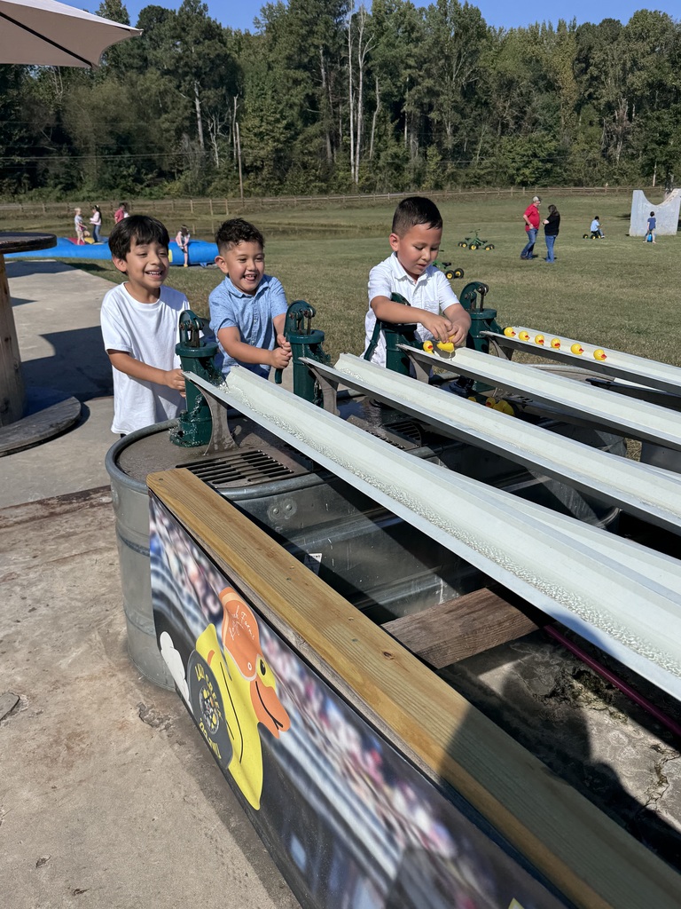 Three elementary students laugh and play at an outdoor rubber duck race station. They pump green hand-operated water pumps to send yellow rubber ducks down metal troughs on a sunny day.