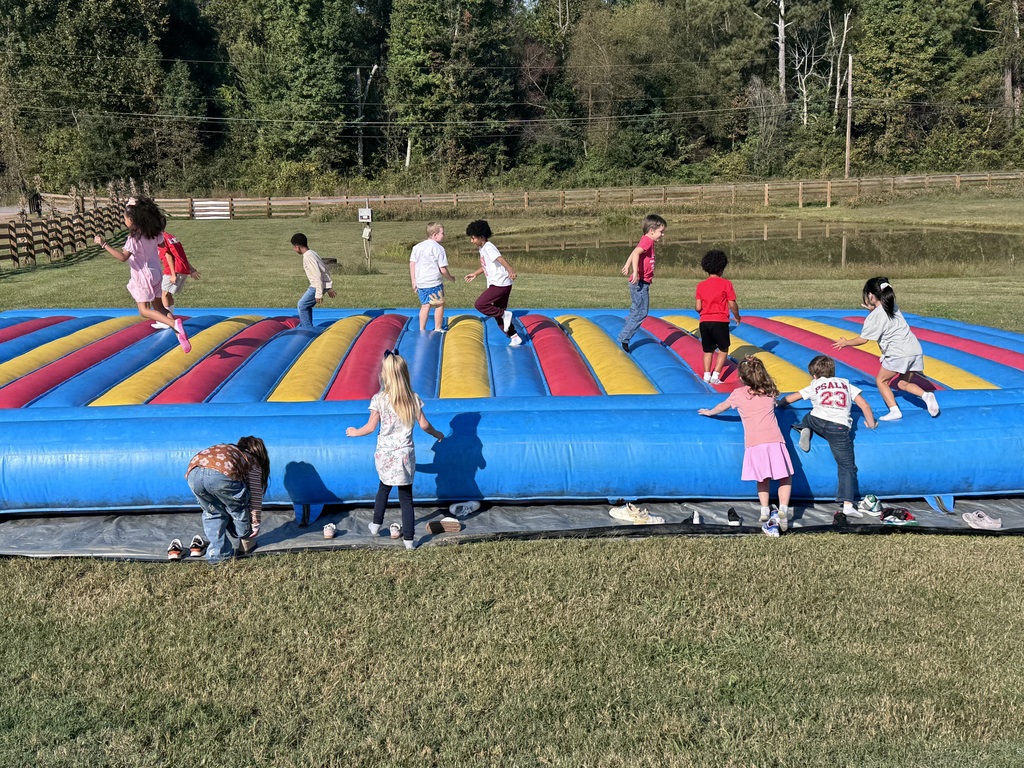 Students happily jump, run, and play on a giant blue, yellow, and red inflatable jumping pad set outdoors in a grassy field. In the background are trees, a wooden fence, and a pond under bright sunlight.