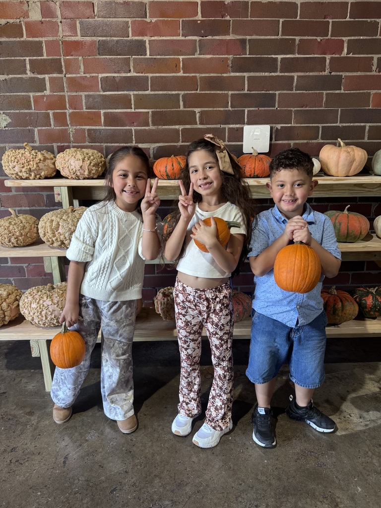 Three young children stand together smiling and making peace signs while holding small orange pumpkins. Behind them are wooden shelves loaded with various pumpkins and gourds against a brick wall