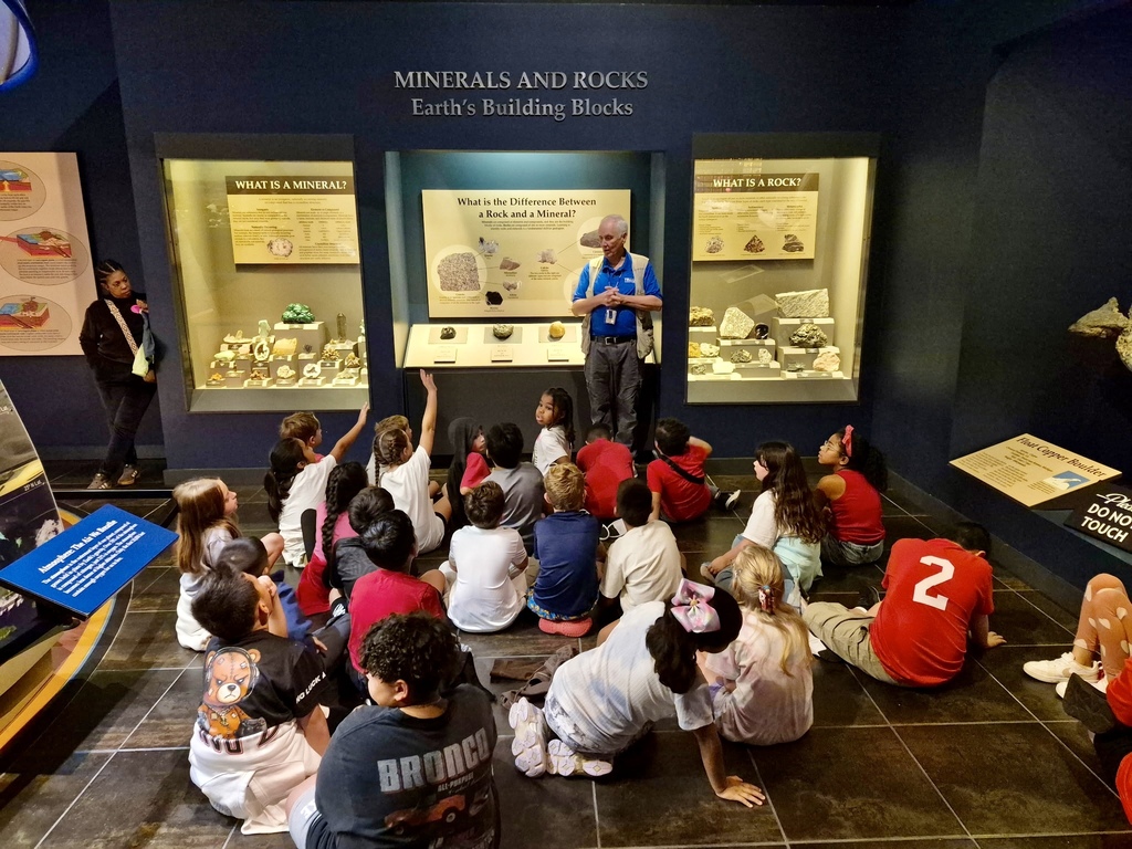 A group of approximately twenty students sitting on a dark tile floor inside a science museum, facing a museum instructor. The instructor, wearing a blue shirt and beige vest, stands in front of a wall display titled "MINERALS AND ROCKS: Earth's Building Blocks" featuring illuminated exhibit cases with rock and mineral specimens. Several students have their hands raised to answer or ask questions during the presentation.