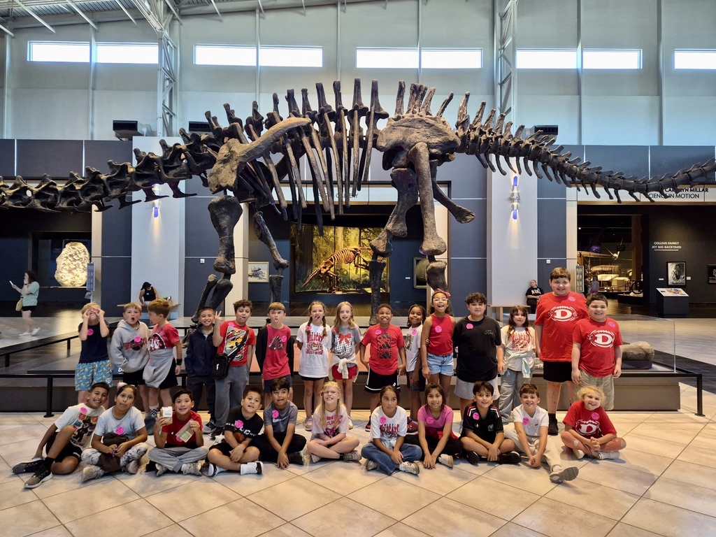 A class of over twenty elementary school students poses together in a spacious, brightly lit museum hall. The students are arranged in two rows—one sitting cross-legged on the tiled floor and the other standing behind them—with many wearing matching pink name tags and red or white school t-shirts. Looming directly behind the entire group is a huge, full-body fossil skeleton of a dinosaur displayed on a raised platform.
