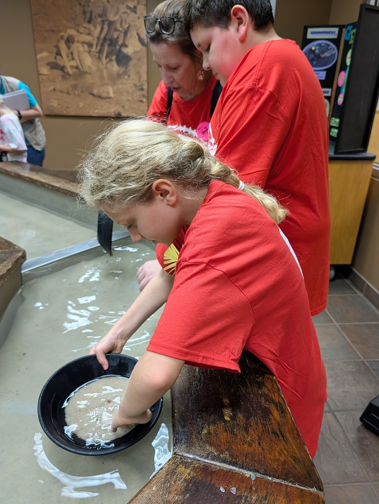 A blonde girl in a red t-shirt stands at a wooden water trough, carefully sifting sand with her hands inside a black panning pan submerged in water. Beside and behind her, another student and an adult chaperone in matching red shirts watch closely as she searches for hidden minerals or fossils.