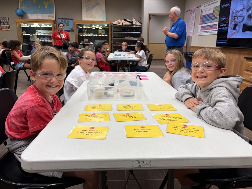 Four young students wearing clear safety goggles smile at the camera while seated at a white activity table in a classroom or lab setting. In the center of the table, bright yellow index cards displaying clues about mineral properties—such as luster, magnetism, and crystal shape—are laid out in rows. Other students and teachers working in the background complete the classroom environment.