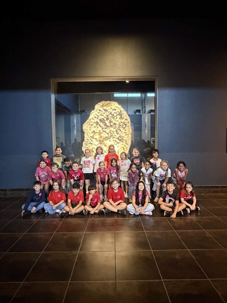 A class of approximately 25 elementary school students poses together for a group photo inside a dark museum gallery. The children are arranged in three rows, sitting cross-legged or kneeling on a dark tiled floor, with many wearing matching pink name tags and red or white school shirts. Directly behind them is a large glass display case framing a huge, brightly lit, crystalline mineral specimen.