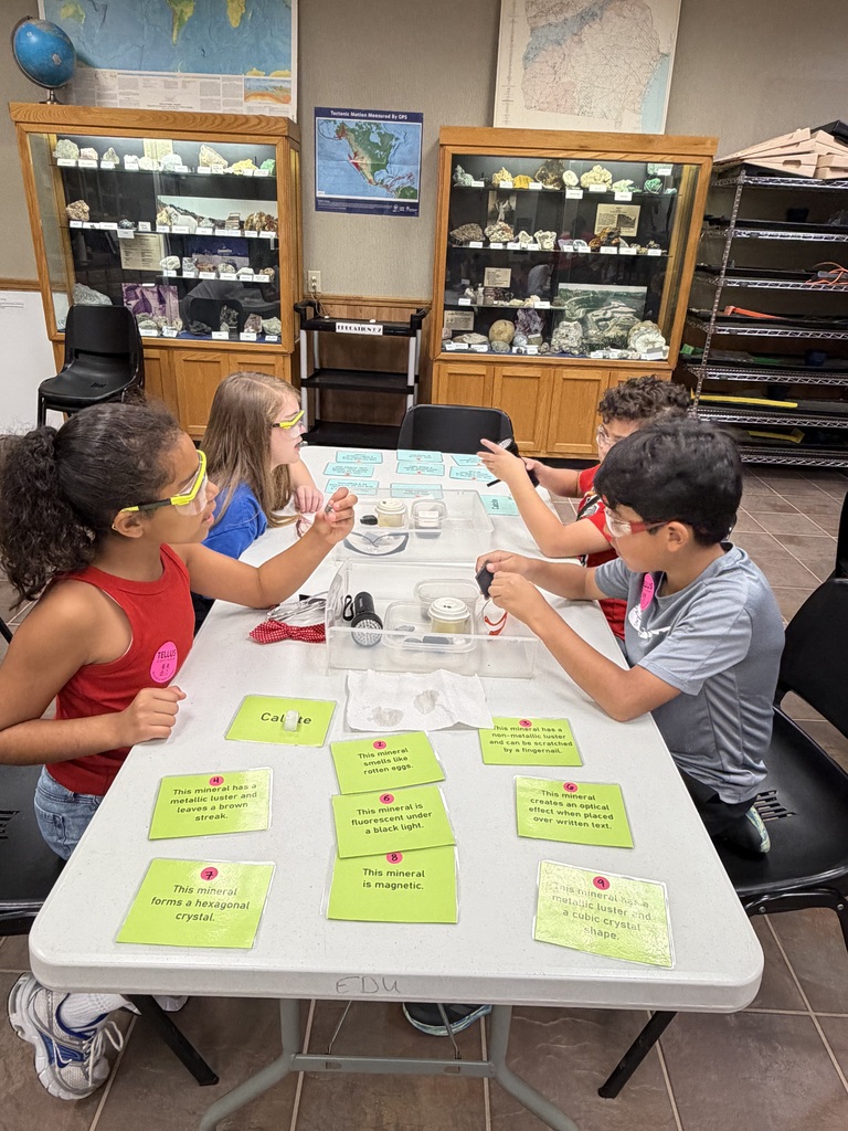 Four young students wearing protective safety glasses gather around a long white activity table in a classroom lab. Yellow activity cards with mineral clues—such as "This mineral is magnetic" and "This mineral forms a hexagonal crystal"—are laid out on the table alongside testing equipment, including small bottles and magnifying tools. Glass display cabinets filled with geological rock and mineral samples are visible in the background.