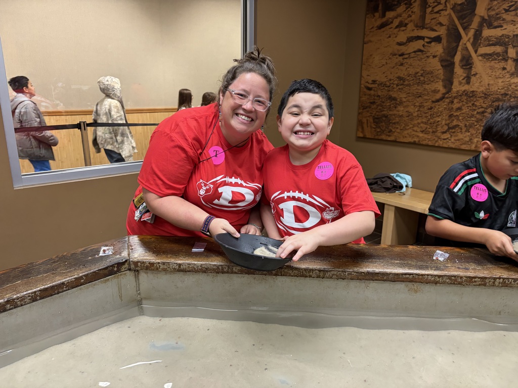 A female teacher wearing glasses and a red school shirt smiles beside a young male student who is grinning brightly. Both are wearing matching red shirts featuring a football and school logo, along with pink name tags. They are holding a black panning pan together over a wooden water trough filled with sand and water in an educational museum setting.