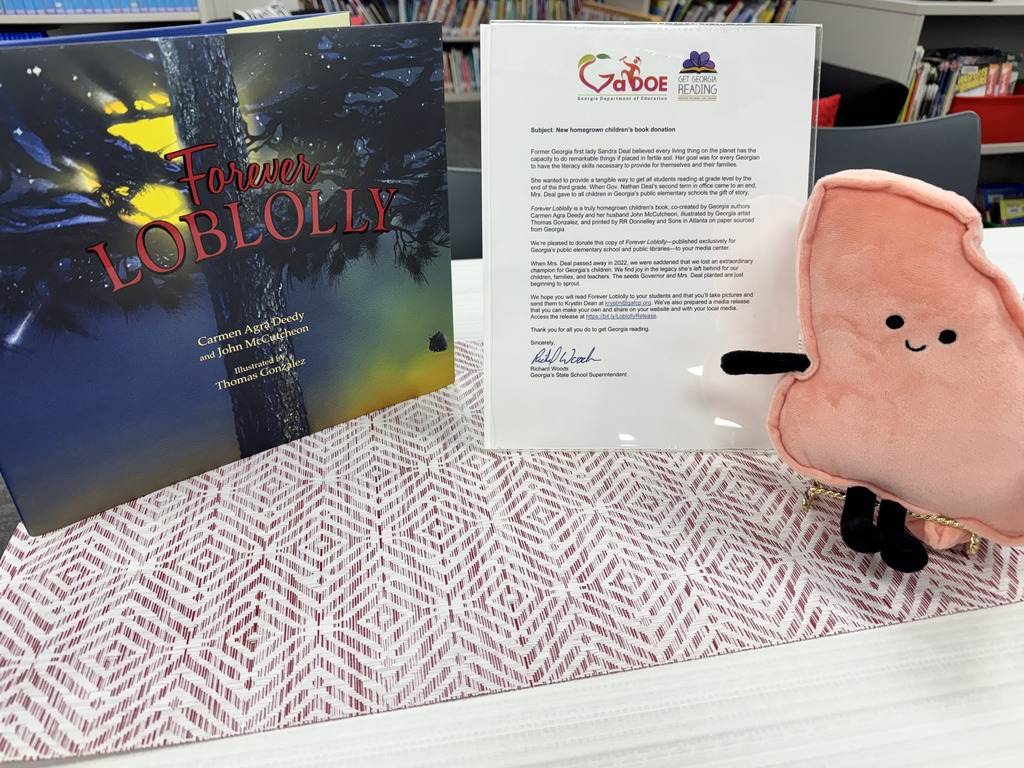 A special book donation to the City Park Elementary Media Center featuring Forever Loblolly, a children's book by Georgia authors Carmen Agra Deedy and John McCutcheon, illustrated by Thomas Gonzalez. The book is displayed alongside a donation letter from Georgia State School Superintendent Richard Woods and a smiling pink book character. The letter explains that the donation honors the literacy legacy of former Georgia First Lady Sandra Deal and her dedication to helping Georgia's children become successful readers.
