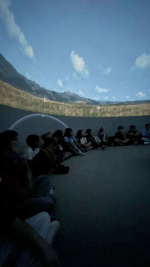 Close-up perspective of elementary students sitting on the floor in a dim planetarium dome, looking up at a projected mountain landscape