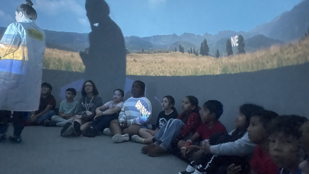 Elementary students sit in a semi-circle on the floor inside a planetarium dome, watching a projected landscape with mountains and a blue sky overhead
