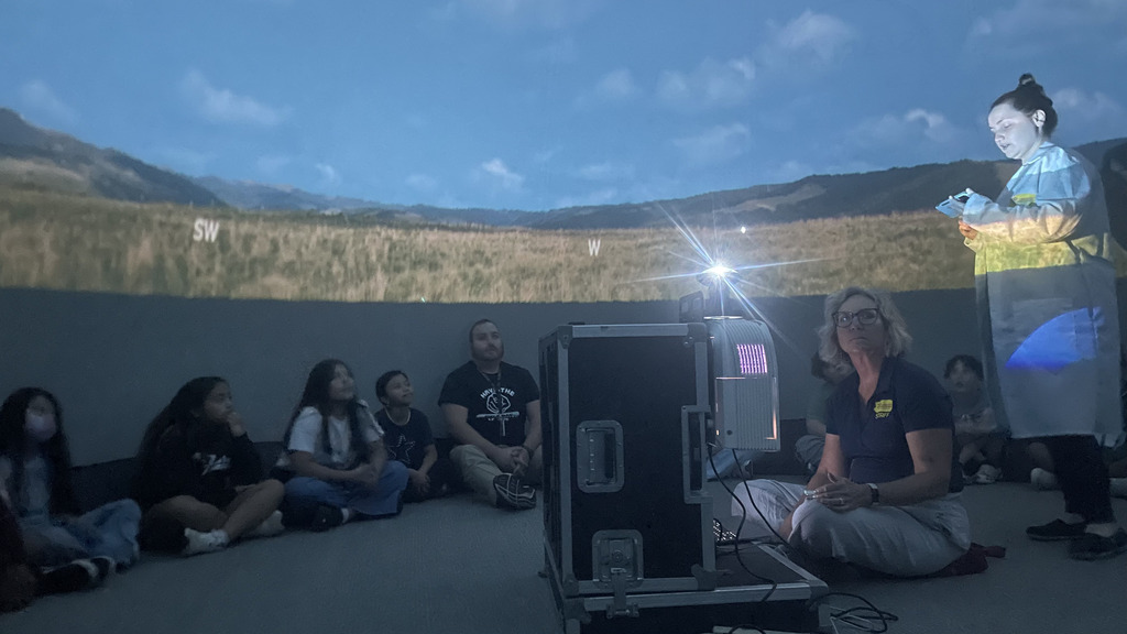 A group of elementary students sitting on the floor inside a planetarium, watching an instructor and a projected projection of a grassy landscape on the curved dome wall