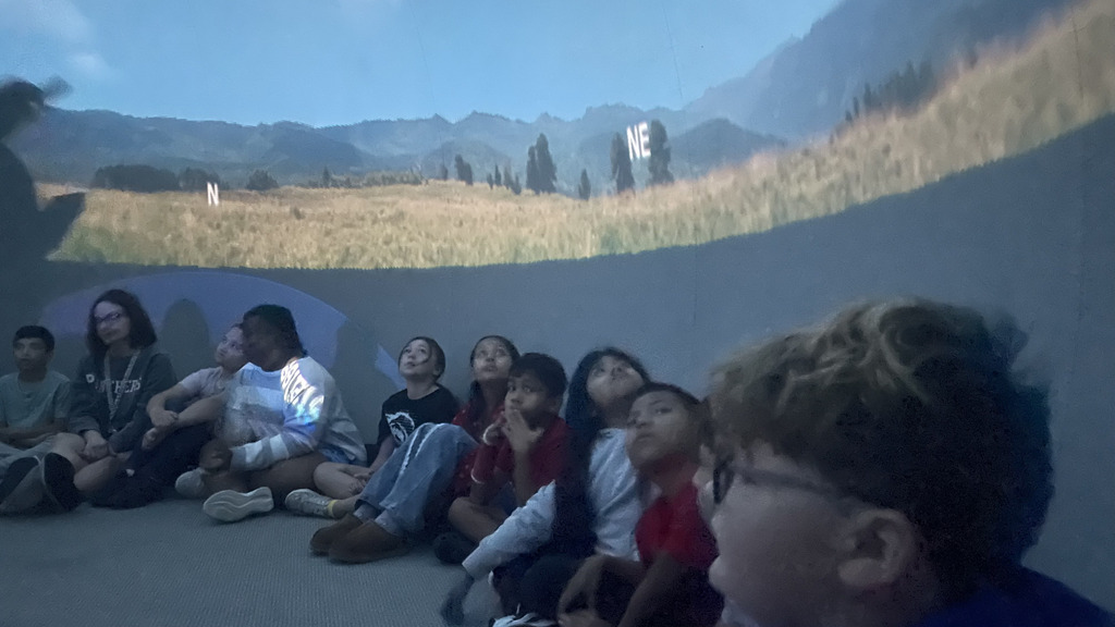 Elementary school students sit on the floor inside a planetarium while an instructor near a projector guides the presentation projected onto the dome screen.