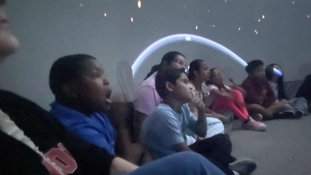 A group of elementary students sitting in a dark planetarium, looking up in awe and excitement at stars projected onto the dome overhead.