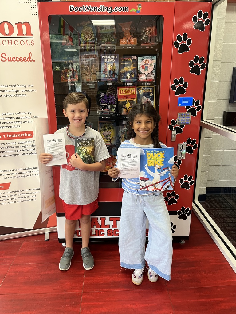 Two smiling students standing together in front of a red book vending machine. The boy on the left holds a Percy Jackson book and paper, while the girl on the right holds a book titled Duck on a Bike along with her award form.