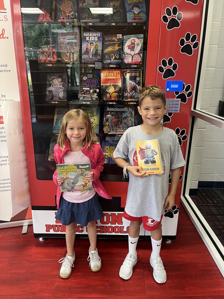 Two students standing side by side in front of a school book vending machine. The girl on the left holds an elephant picture book, and the boy on the right holds a Gordon Korman chapter book titled Slacker.