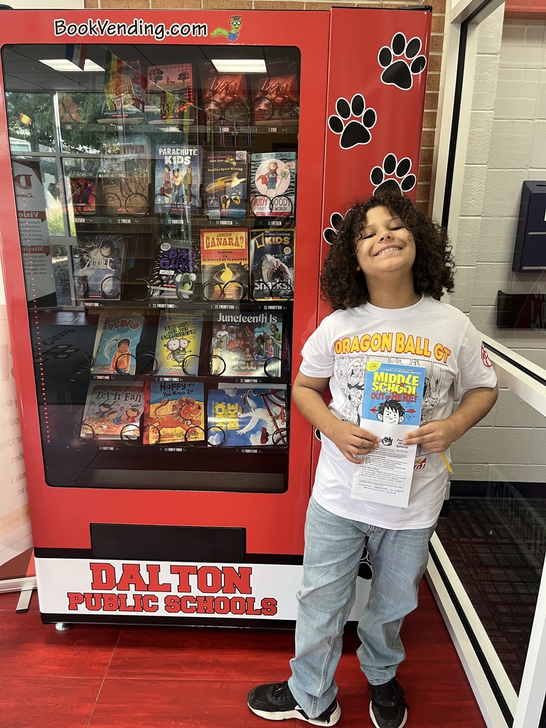 A smiling student with curly hair standing next to a red book vending machine, holding up a paper referral form and a copy of the book Middle School: Get Me Out of Here!.