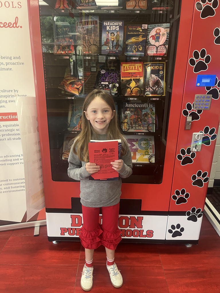 A young student smiling in front of a red book vending machine, wearing red pants and a grey top, while holding a red Positive Office Referral form and a book.