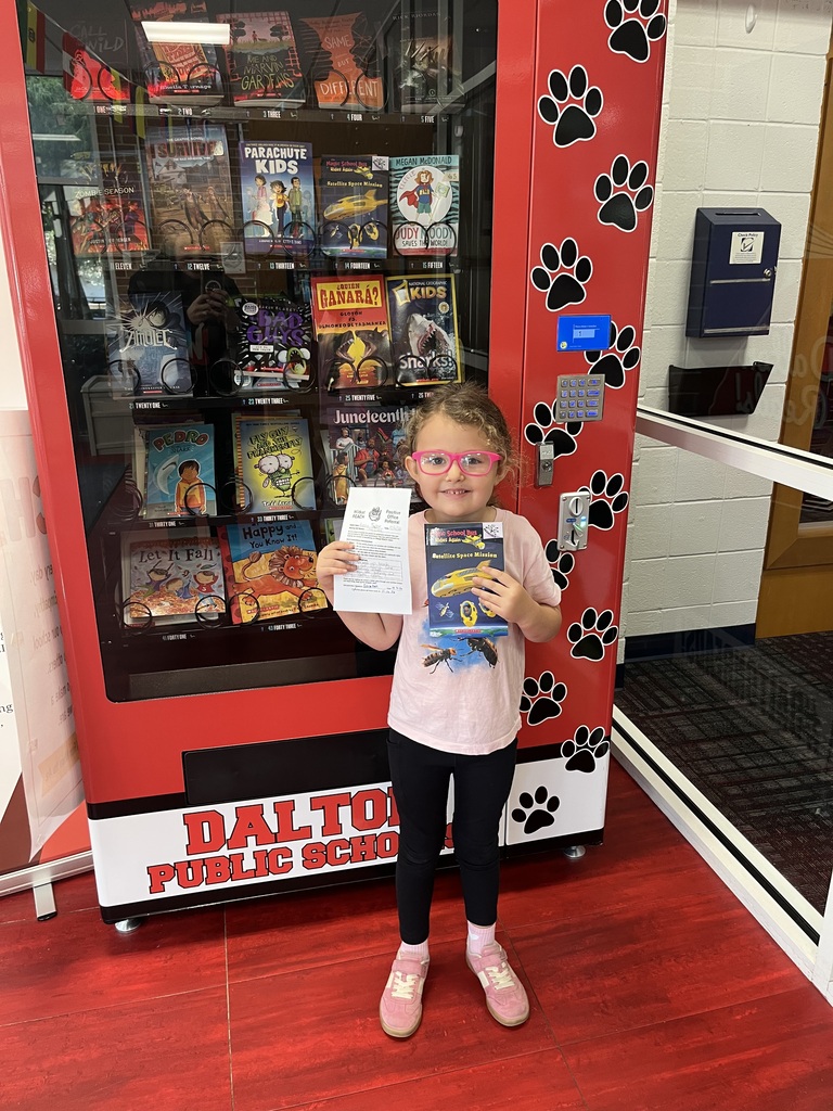 A young girl with bright pink glasses standing in front of a red Dalton Public Schools book vending machine, smiling as she holds a book titled Satellite Space Mission and a paper referral form.
