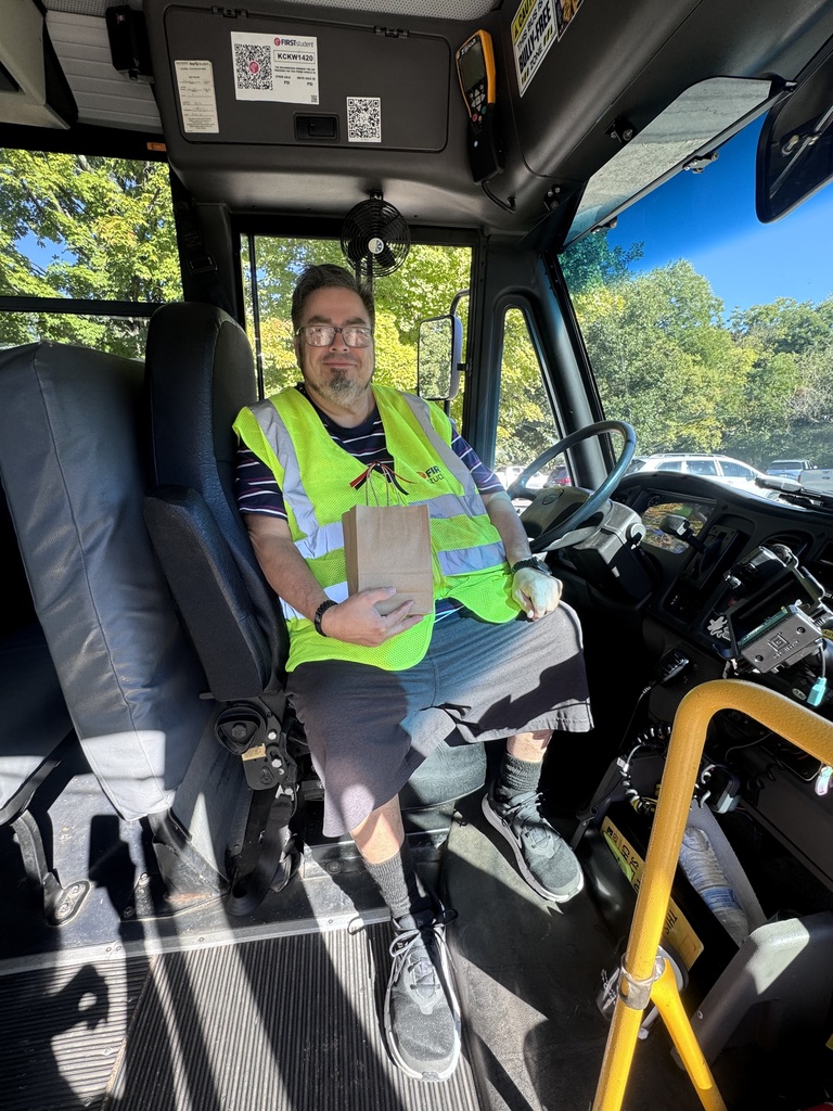 A smiling school bus driver with glasses and a goatee sits in the driver's seat inside a bus. He is wearing a neon green safety vest over a striped shirt and holding a small brown paper gift bag with both hands. Sunny trees and park vehicles are visible through the bus windows.
