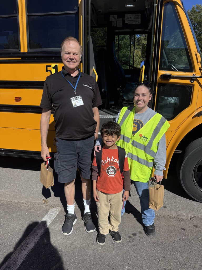 A young boy wearing a red shirt and backpack stands smiling in front of an open yellow school bus door. He is flanked by a man on the left in a black polo shirt and lanyard, and a woman on the right in a high-visibility safety vest. Both adults are smiling and holding small brown paper gift bags outside on a sunny day.