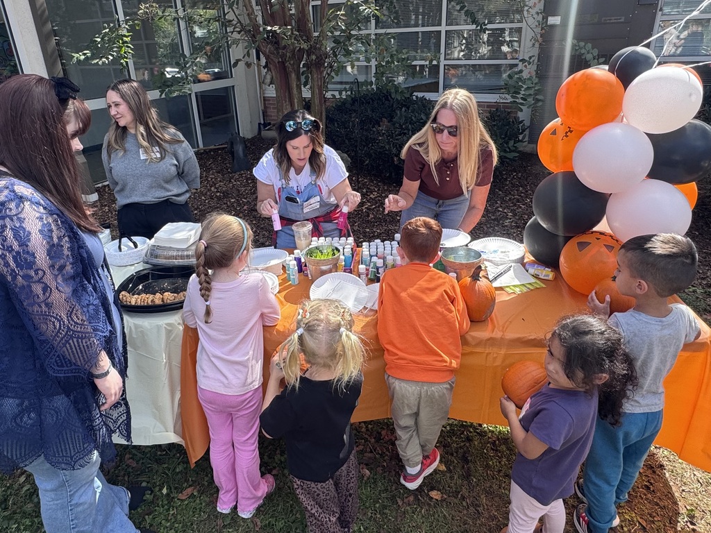 Adult volunteers and children gathered around a outdoor activity table covered with an orange tablecloth. On the table, paint bottles, cups, and paper plates are set up alongside trays of snacks and an orange, black, and white balloon arch. Two women behind the table assist young students in choosing paint bottles, while parents stand nearby watching as the children prepare to decorate small pumpkins.